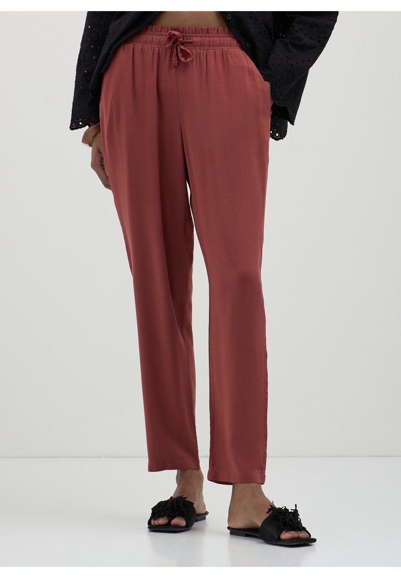 Personne portant un pantalon à cordon de couleur rouille et des sandales noires ouvertes à l'avant avec des nœuds texturés, debout sur un fond uni.