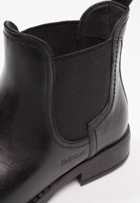 Botín negro de cuero hasta el tobillo con panel lateral elástico, suela texturizada y detalle de marca "GEOX" en el lado exterior.