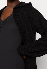 Cárdigan negro de punto con capucha, que presenta un patrón texturizado y mangas largas, llevado sobre una camisola negra ajustada con borde de encaje.