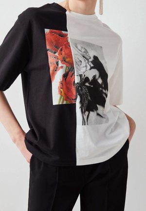 Osoba nosi pół czarną, pół białą koszulkę z czerwonymi i szaro-skalowymi kwiatowymi nadrukami podzielonymi na pół, w połączeniu z czarnymi spodniami i wiszącymi kolczykami.