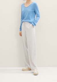 Frau trägt einen hellblauen V-Ausschnitt-Pullover, hochtaillierte, weite weiße Hosen und cremefarbene Slipper und steht mit einer Hand in der Tasche.