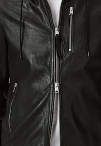 Veste en cuir noir avec une texture lisse, plusieurs zips en argent et des cordons de serrage ajustables sur la capuche. Vue rapprochée des détails du design.