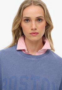 Sudadera azul con impresión de "BOSTON", combinada con una camisa de cuello rosa. Tela suave, corte relajado, escote redondo, con costuras visibles.