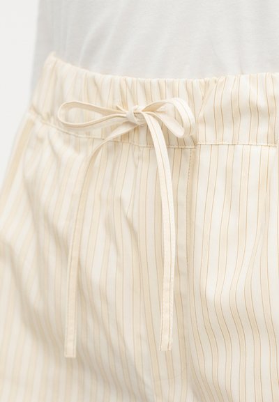 Περιοχή μέσης κρεμ παντελονιών με λεπτές κάθετες ρίγες και κορδόνι δεμένο σε φιόγκο, φορεμένο με λευκό τοπ που είναι μπουκαρισμένο μέσα.
