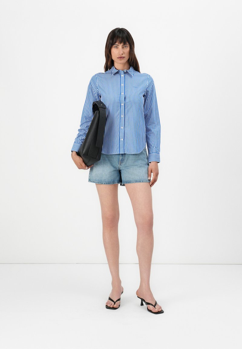 Blau-weißer gestreifter Button-up-Hemd, Jeansshorts, schwarze Handtasche und schwarze Absatzflip-flops. Schlichter Hintergrund, Model steht aufrecht.