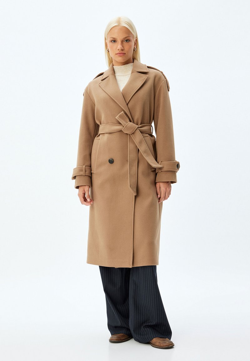 Kamelfärgad trench coat med bältesmidja, skräddarsydda slag och stora framknappar. Texturerad tyg, långa ärmar och sidfickor.