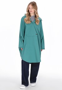 Poncho impermeabil turcoaz cu glugă, mâneci lungi și buzunar frontal. Poartă peste un tricou în dungi, asortat cu blugi largi de culoare închisă și pantofi albi.