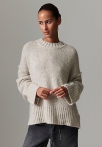 Camisola de malha bege clara com barra canelada e mangas largas. Textura suave com corte relaxado e decote redondo. Combinada com jeans escuros.