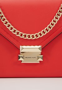 Rød læder skuldertaske med en guldkæde-rem, geometrisk lås og præget logo. Glat tekstur med en struktureret form.