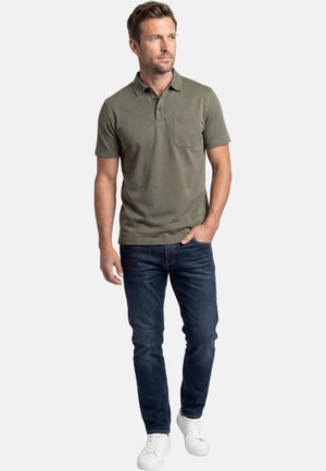 Man staat en draagt een olijfgroen poloshirt met korte mouwen en zakje, donkerblauwe spijkerbroek en witte sneakers, kijkt naar rechts.