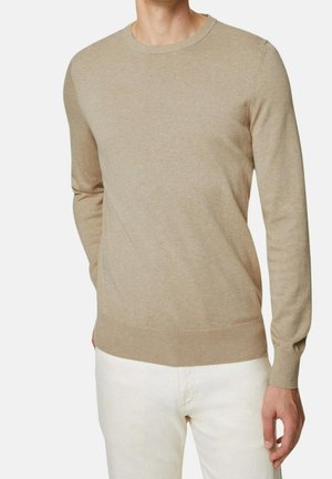 Homme portant un pull en maille beige à manches longues et un pantalon blanc, debout avec une main dans la poche contre un fond uni.