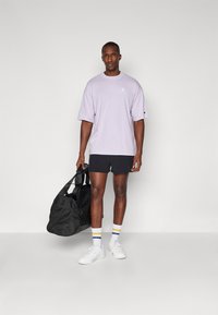 Lavendel bomulds t-shirt med korte ærmer, sorte shorts og hvide sneakers. Holder en sort sportsbag, iført stribede strømpers.