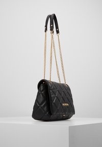 Borsa a tracolla in pelle trapuntata nera con catene dorate e un profilo sottile. Presenta un accento con logo sulla parte anteriore e una chiusura a patta.