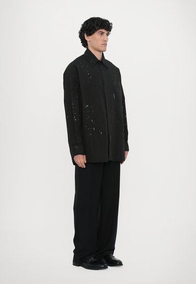 Chemise noire à boutons, au fit oversize, présentant des motifs éclaboussés en bleu clair. Associée à un pantalon noir plissé et des chaussures noires brillantes.