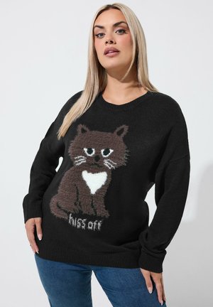 Vrouw met lang blond haar die een zwarte trui draagt met een bruine kat met een witte borst en de woorden "hiss off" en een blauwe spijkerbroek.