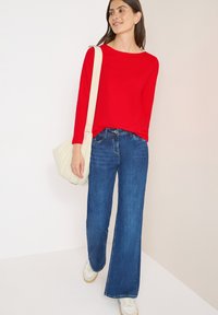 Maglietta rossa a maniche lunghe, jeans a gamba larga blu e sneakers bianche. Borsa a spalla beige. Tessuti morbidi e vestibilità rilassata per un look casual.