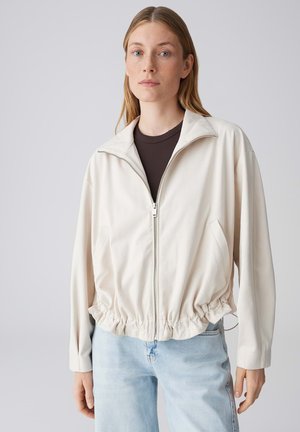 Giovane donna che indossa una giacca beige chiaro con zip sopra una maglia scura e jeans azzurri, in piedi davanti a uno sfondo grigio uniforme.