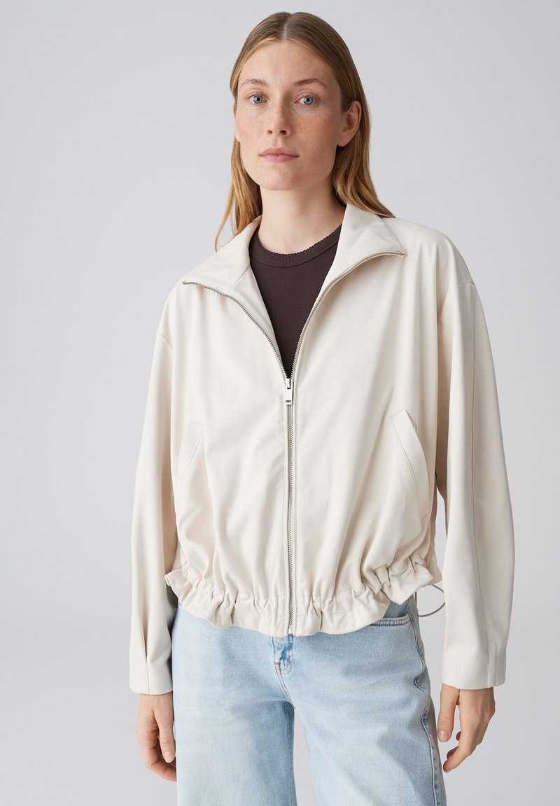 Jeune femme portant une veste zippée beige clair sur un haut foncé et un jean bleu clair, debout devant un fond gris uni.