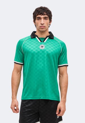 Mężczyzna ubrany w zieloną kratowaną krótką sportową koszulkę z czarnym kołnierzem i pasujące czarne szorty, stojący na jednolitym tle.