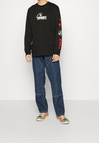 Camisa negra de manga larga con diseños gráficos en blanco y rojo; combinada con pantalones de mezclilla azules y zapatillas naranja y grises.