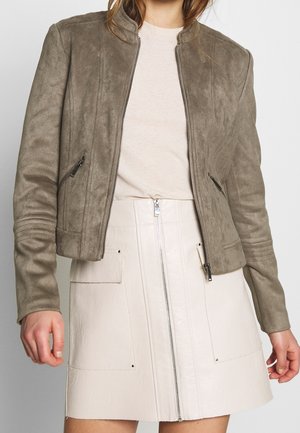 Femme portant une veste zippée en daim taupe sur un haut beige clair et une jupe crème taille haute avec fermeture éclair sur le devant et grandes poches.