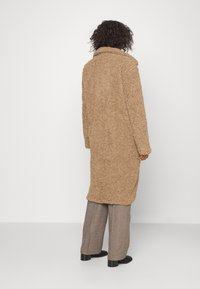 Persona con capelli ricci che indossa un lungo cappotto beige strutturato, pantaloni grigi a gamba larga e scarpe nere, in piedi contro uno sfondo bianco.