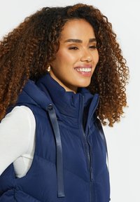 Smilende kvinde med krøllet hår iført guldringe i ørerne og en marineblå dunvest over en hvid langærmet skjorte mod en hvid baggrund.