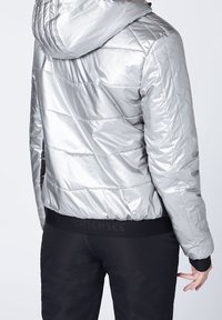 Silver puffjacka med huva, med ett quiltat mönster och elastisk midja. Bärs med svarta byxor som visar en snygg, glänsande textur.