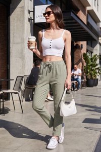 Mujer con gafas de sol, camiseta corta blanca y pantalones verdes sostiene una taza de café y un bolso blanco mientras camina fuera de una cafetería.