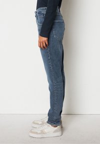 Modré denimové džíny se rovnými nohavicemi, vysokým pasem a jemným opotřebením. Spárované s bílými teniskami se béžovými akcenty.