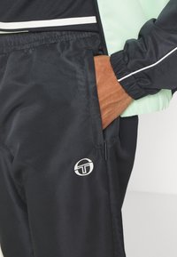 Juodos sportinės kelnės su tekstūruotu paviršiumi, turinčios šoną užtraukiamą kišenę, elastingą juosmenį ir apvalią baltą logotipą ant kojos.