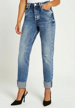 Femme portant un jean en denim bleu clair retroussé avec une petite déchirure à la cuisse, assorti à des escarpins noirs pointus à bride arrière.