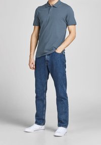 Šedá polo košile vyrobená z bavlny, zkombinovaná s modrými džíny. Oba kusy mají uvolněný střih. Celé outfit doplňují bílé tenisky. Na košili je minimální branding.