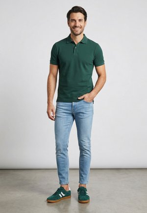 Mann steht drinnen, trägt ein dunkelgrünes Poloshirt, hellblaue Jeans und grüne Sneakers mit weißen Streifen, lächelt in die Kamera.