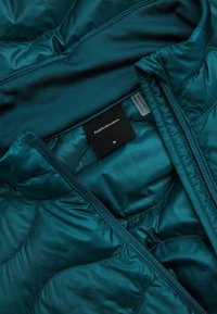 Giacca trapuntata turchese, parzialmente aperta, con etichetta nera del marchio PeakPerformance e taglia "M" all’interno del colletto.