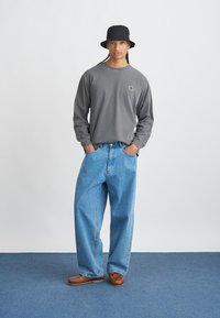Hallikas pikkade varrukatega särk logo kanssa, helesinised laiad teksad, must bucket hat ja pruunid mokassinikujulised jalanõud tavalise tausta ees.