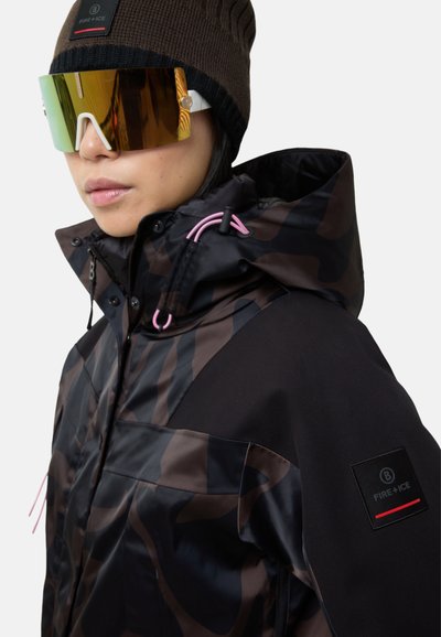 Braune und schwarze Tarnjacke mit hohem Kragen, versehen mit pinken Akzentzugbändern und einem Logopatch am Ärmel. Große reflektierende Sonnenbrille.