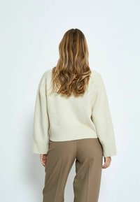 Maglione oversize color crema con polsini e orlo a coste, indossato con pantaloni marroni a vita alta e taglio sartoriale. Tessuto morbido, vestibilità rilassata.