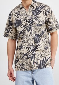 Beżowa koszula z krótkim rękawem z czarnym nadrukiem w kwiaty, zapinana na guziki z przodu, z dwiema kieszeniami na piersi i małą metką z logo na końcu.