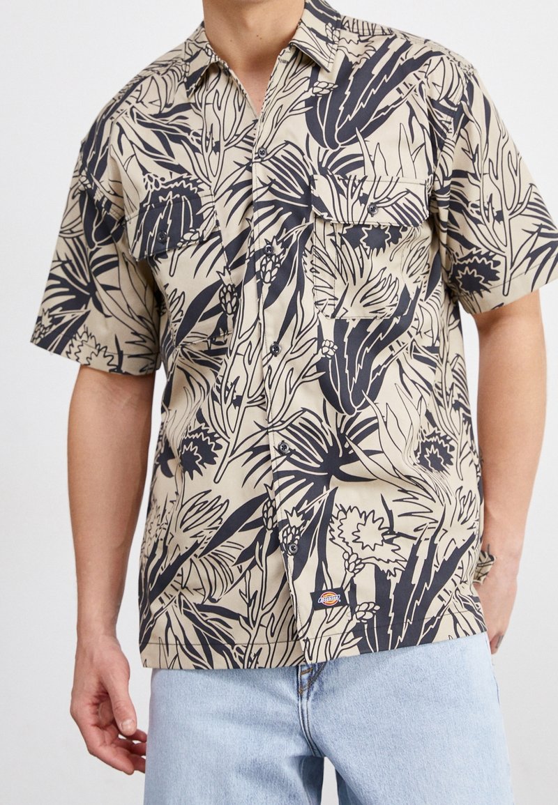 Beżowa koszula z krótkim rękawem z czarnym nadrukiem w kwiaty, zapinana na guziki z przodu, z dwiema kieszeniami na piersi i małą metką z logo na końcu.