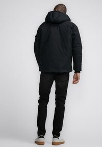 Schwarze gepolsterte Jacke mit Kapuze, die eine glatte Oberfläche und lange Ärmel hat, kombiniert mit schwarzen Jeans und weißen Sneakers.