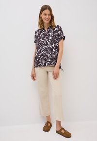 Chemise à manches courtes avec imprimé feuillage noir et blanc, associée à un pantalon large beige et des sabots en suède marron. Design simple avec des poignets retroussés.