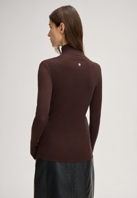 Brauner, gerippter Rollkragenpullover mit langen Ärmeln und einem kleinen Metallelement auf der Rückseite. Er hat eine figurnahe Passform und einen strukturierten Stoff.