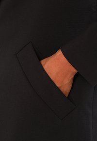 Black garment with a sleek, smooth texture. Features a side pocket with a rectangular opening, showcasing a hand resting inside.