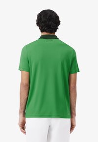 Grön pikétröja med krage, bakvy; har en slät yta och Lacoste-logotyp på nacken. Bärs med vita shorts.