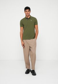 Olivově zelené polo tričko s límečkem a krátkými rukávy, kombinované s béžovými kalhotami s širokými nohavicemi a černými botami. Jednoduchý design s logem.