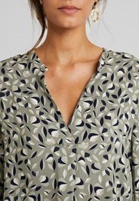 Top verde com padrão, apresentando um decote em V, com designs abstratos de folhas em tons de azul marinho, off-white e cinza. Tecido leve e suave.