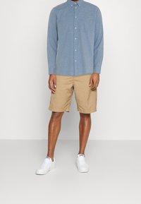 Chemise à manches longues bleu clair avec un poche poitrine, associée à un short beige jusqu'aux genoux et des sneakers blanches.