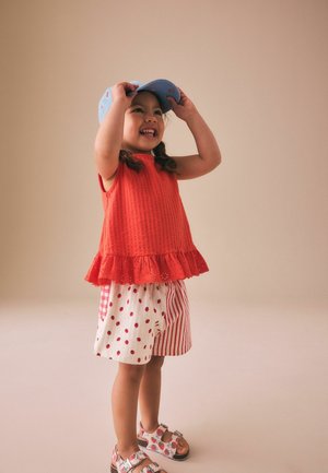Smilende lille pige i rød ærmeløs top, mønstrede shorts og sandaler justerer en blå kasket mod en ensfarvet beige baggrund.