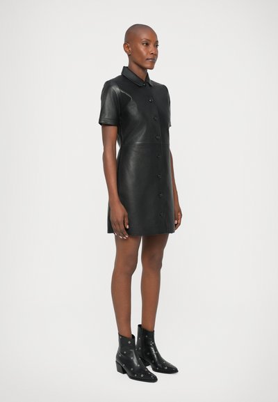 Czarna skórzana sukienka koszulowa z krótkimi rękawami, zapięciem na guziki i kołnierzykiem. Noszona z czarnymi botkami za kostkę z ozdobami w postaci srebrnych okuć.